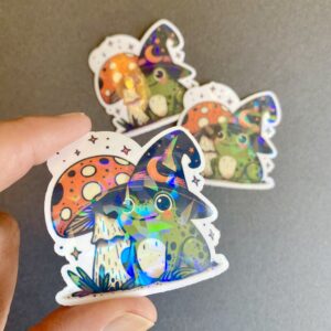 adorable frog wearing a witches hat and sitting by a mushroom, holographic vinyl die-cut sticker