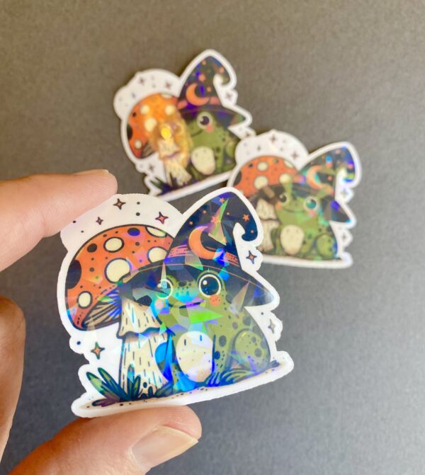 adorable frog wearing a witches hat and sitting by a mushroom, holographic vinyl die-cut sticker